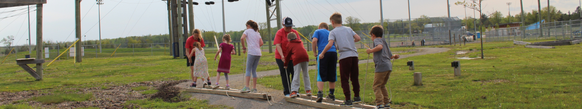 A group of children standing in a line on a wooden beam outdoors, balancing or walking along the beam as part of an activity. The background includes grassy areas, trees, and structures suggesting an outdoor adventure or ropes course area.